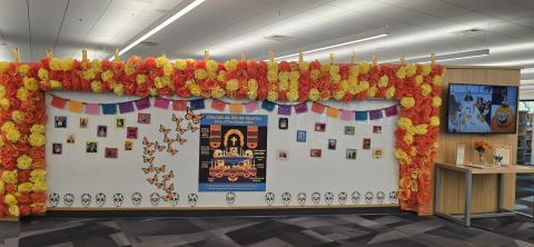 Marigold paper tissue flowers surrounding the North Entrance Arc Wall. LED Candles placed on top. A poster of what is commonly found on a Day of the Dead altar. Monarch butterflies, paper sugar skulls, paper banner adorn the wall. Pictures of loved ones shared by community members.