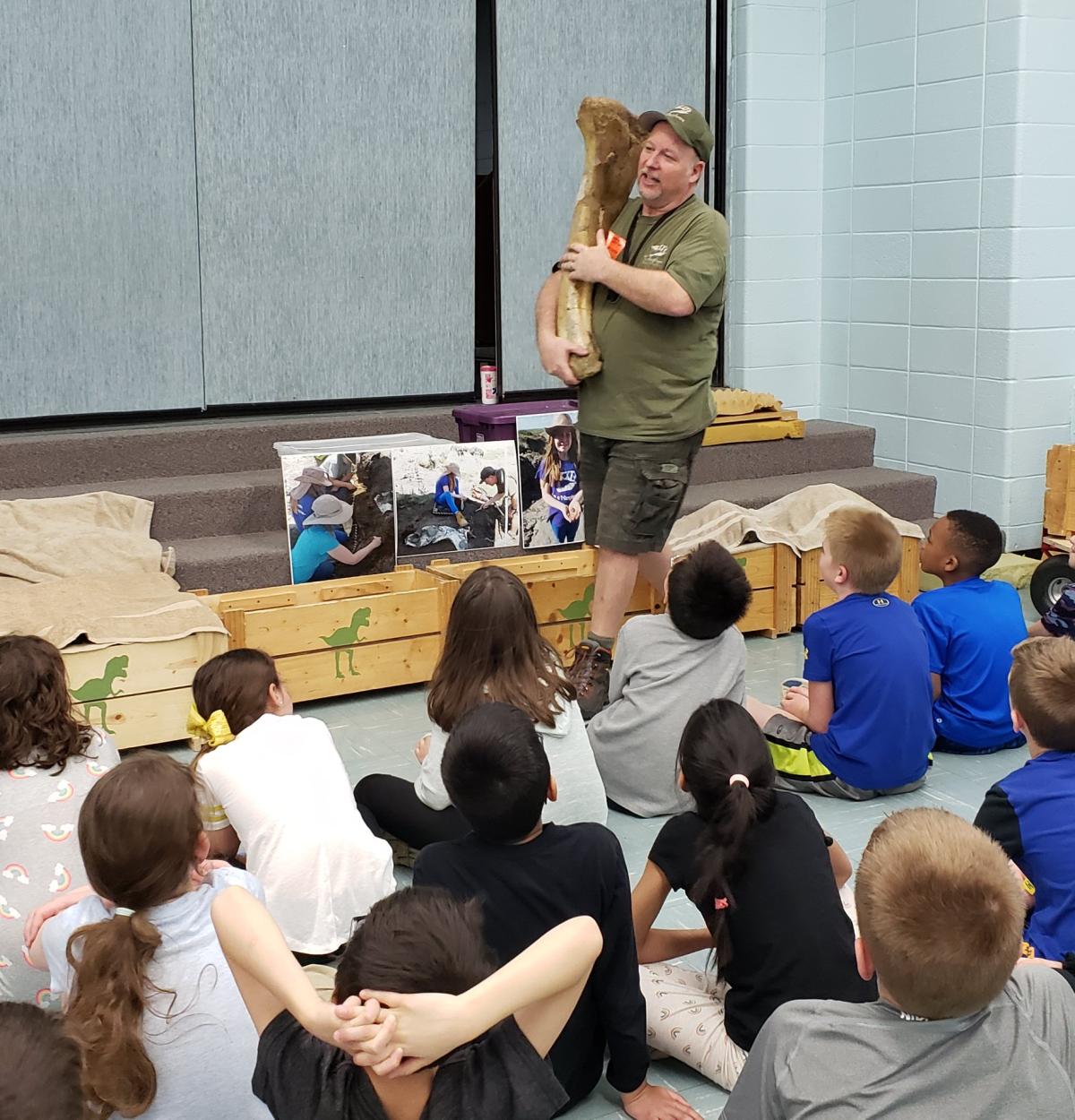 David of the T-rexplorers holding a large dinosaur fossil in front of a room of children.